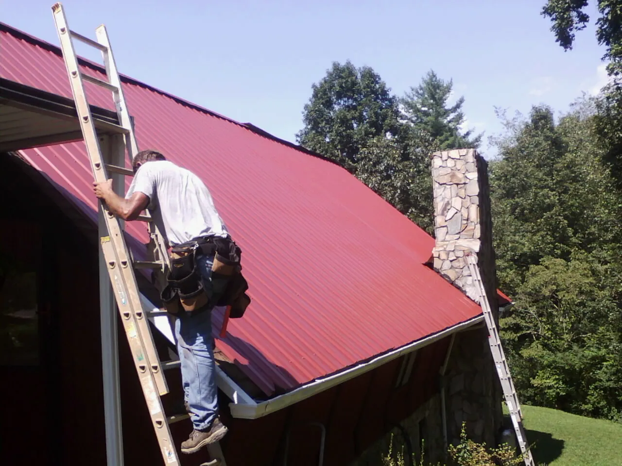 Roofer on a ladder beside a red metal roof and stone chimney — Soffit & Fascia Repair by Roofing Brothers in Foosland
